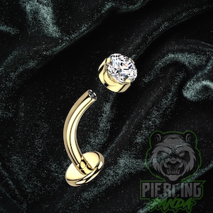 May include: A gold-colored curved piercing jewelry with a clear, round gemstone. The jewelry is designed with a curved bar and a circular base. The gemstone is set in a gold-colored bezel.