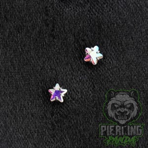 May include: Two star-shaped jewelry studs featuring iridescent, multi-colored crystal centers set in silver-toned metal prongs. The top star displays clear and pinkish hues, while the bottom star shows a deep purple center with iridescent edges.