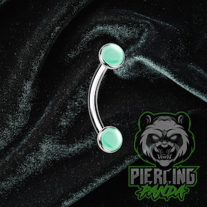 May include: Curved eyebrow piercing jewelry with a polished silver-tone bar and two light green, round, cabochon-style ends. The jewelry is set against a dark, textured background. The logo "Piercing Panda" is visible.