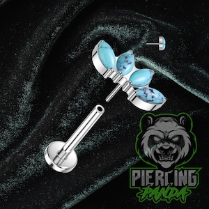 Puede incluir: Un piercing labial plateado con una parte superior decorativa con tres piedras de color turquesa con vetas azul oscuro. También se muestra un pequeño perno a juego. El fondo es de terciopelo verde oscuro.