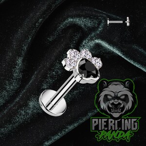 May include: A silver-colored labret stud with a flower-shaped design. The flower has five clear, round gems surrounding a black heart-shaped gem. The stud has a flat, round base and a straight post.