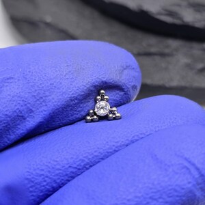 May include: A silver triangle-shaped body jewellery piece with a clear gemstone in the centre. The piece is made up of small silver balls.