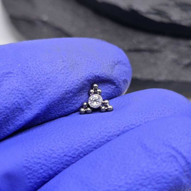 May include: A silver triangle-shaped body jewellery piece with a clear gemstone in the centre. The piece is made up of small silver balls.