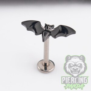 May include: A black bat-shaped stud earring with a metallic post and a flat circular base. The bat design features smooth, glossy wings and a small head, attached to a straight metal stem. The overall construction is a single piece of jewelry.