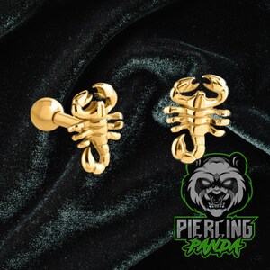 May include: A pair of gold-colored scorpion-shaped earrings. The earrings are detailed with a curved tail and multiple legs. The background is a dark, textured fabric. The logo "Piercing Panda" is visible in the bottom right corner.