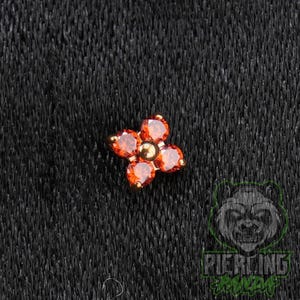 May include: A flower-shaped body jewelry stud featuring four round, faceted orange gemstones arranged around a central gold-toned metal bead. The piece has a metallic gold-colored setting that holds each stone securely in a four-petal floral design.