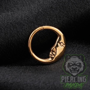 May include: A gold-colored circular hoop ring featuring a raised, embossed design of a human face in profile on one side. The jewelry piece has a smooth, polished metallic finish and a hinged closure mechanism visible at the top of the hoop.