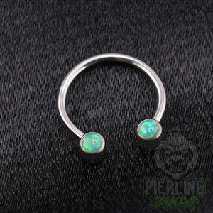 May include: A circular horseshoe-shaped body jewelry piece made of polished silver-toned metal. The two ends of the open ring feature smooth, rounded cabochon beads with a vibrant, iridescent green and blue opal-like finish.
