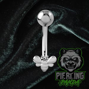 Pode incluir: Um piercing em estilo barbell prateado com um pingente em forma de abelha. O piercing tem uma bola arredondada em uma extremidade e um design de abelha na outra. O fundo é um tecido escuro e texturizado.