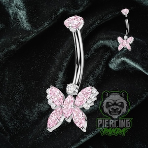 Pode incluir: Um piercing de umbigo prateado com um design de barra curva. O anel apresenta uma grande pedra rosa na parte superior e um design de borboleta com pedras rosas e transparentes na parte inferior. Um anel de borboleta menor também é mostrado.