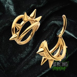 May include: Two gold-colored, spiked earrings. One is a hoop design, and the other is a curved design. The earrings have a shiny, metallic finish and are set against a dark green, velvet-like background.