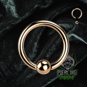 May include: A gold-colored captive bead ring, also known as a ball closure ring, with a polished finish. The ring features a circular design with a small, round bead that secures the ring. A smaller version of the ring is also visible.