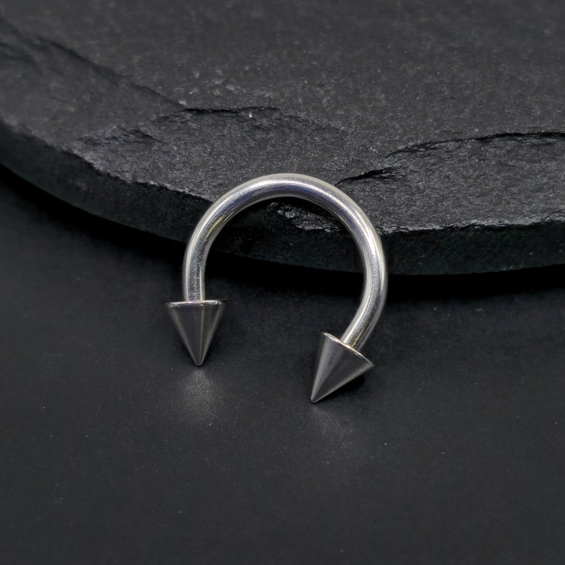 May include: A silver horseshoe piercing with pointed ends. The curved bar has two conical tips. The jewellery is made of a shiny metal and is set against a dark, textured background.