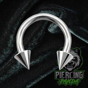 May include: A silver horseshoe piercing with pointed conical ends. The jewellery is set against a dark green velvet backdrop. The logo "Piercing Panda" is visible in the lower right corner.
