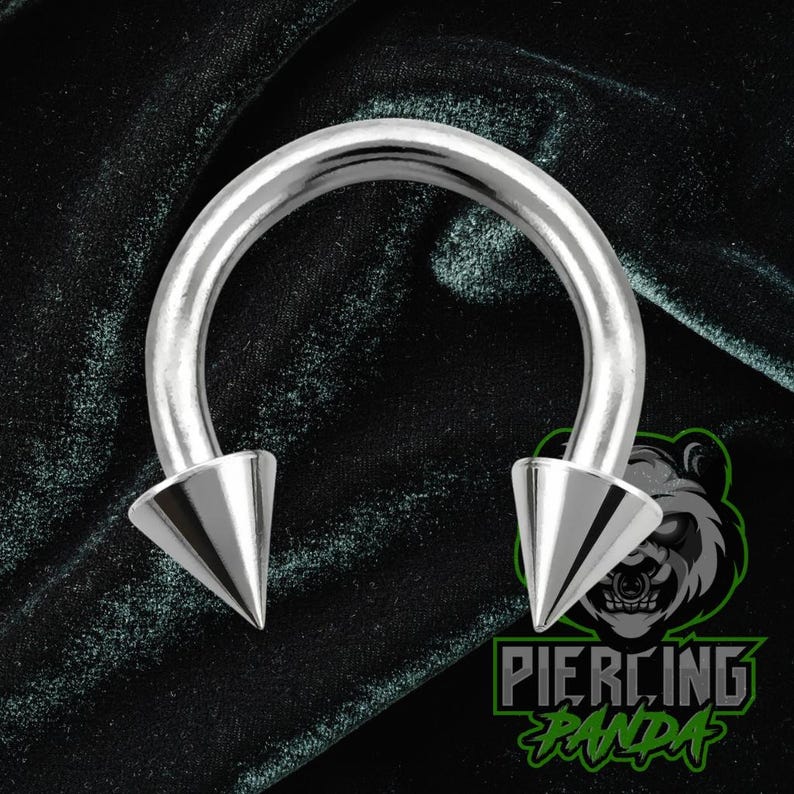 May include: A silver horseshoe piercing with pointed conical ends. The jewellery is set against a dark green velvet backdrop. The logo "Piercing Panda" is visible in the lower right corner.