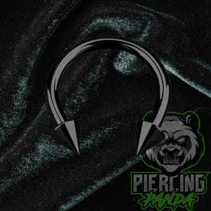 May include: A black circular barbell body jewelry piece featuring two sharp, conical spike ends. The jewelry has a smooth, polished finish and a curved, horseshoe-shaped bar. The design is simple, metallic, and features a dark, monochromatic aesthetic.