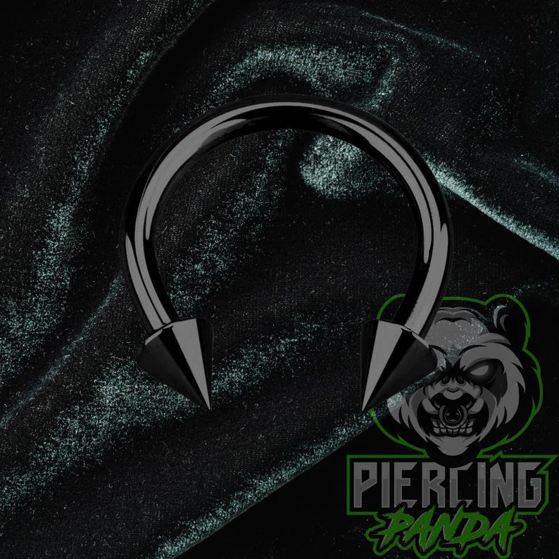 May include: A black circular barbell body jewelry piece featuring two sharp, conical spike ends. The jewelry has a smooth, polished finish and a curved, horseshoe-shaped bar. The design is simple, metallic, and features a dark, monochromatic aesthetic.