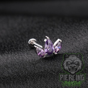 May include: A silver-toned labret stud featuring a decorative top with three purple pear-shaped gemstones arranged in a fan-like cluster. The central stone is the largest, flanked by two smaller stones, all set in a metallic frame with a post.
