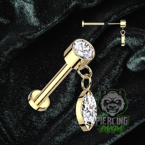 Puede incluir: Tachuela labret dorada con una gema transparente de talla redonda y una gema colgante en forma de marquesa. La joya se muestra sobre un fondo verde oscuro. Se muestran piezas adicionales encima de la tachuela principal.