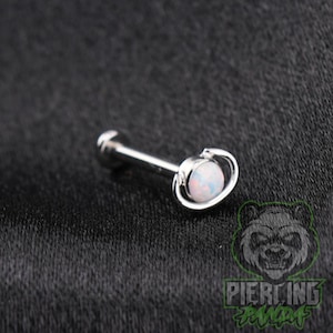 May include: A silver-toned barbell body jewelry piece featuring a round, iridescent white opal-style stone set within a circular frame that resembles the planet Saturn, with a flat circular backing disc at the opposite end of the metal post.