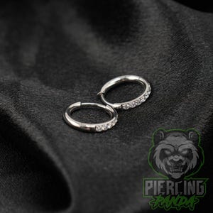 May include: A pair of silver-toned hoop earrings featuring a row of small, clear, sparkling stones embedded along the front curve of each circular band. The earrings have a polished metallic finish and a hinged closure mechanism for secure wear.