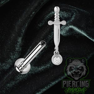 May include: A silver-toned metal body jewelry piece featuring a flat-back labret post and a sword-shaped charm attachment. The sword charm is accented with a single round clear gemstone at the tip. The piece has a polished, reflective finish.