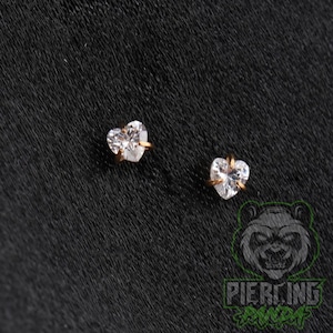 May include: A pair of stud earrings featuring clear, heart-shaped faceted stones set in gold-toned metal prongs. The earrings are displayed against a dark, textured background, showcasing the reflective quality of the stones and the metallic setting.