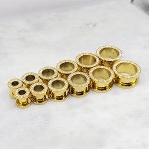 May include: A collection of gold-colored ear gauges in various sizes. Each gauge features a ring of clear, sparkling crystals around the outer edge. The gauges are arranged on a white marble surface.