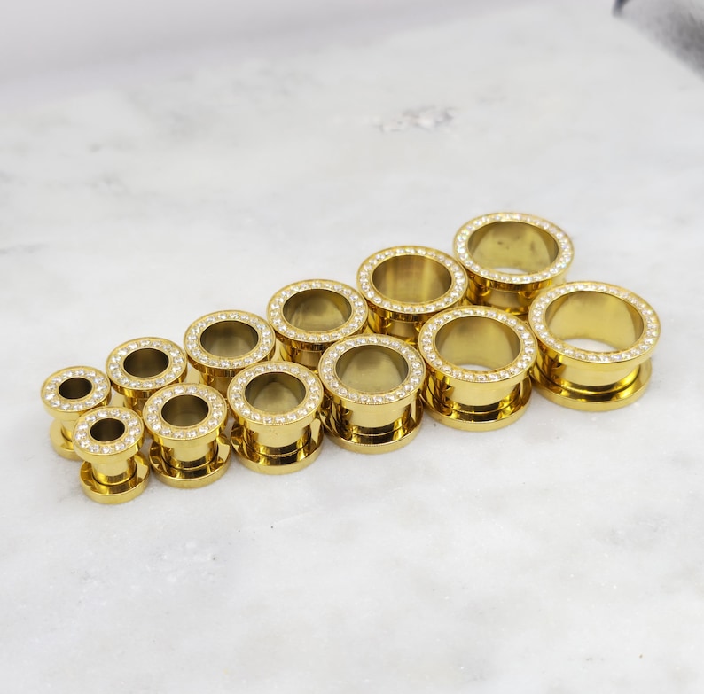 May include: A collection of gold-colored ear gauges in various sizes. Each gauge features a ring of clear, sparkling crystals around the outer edge. The gauges are arranged on a white marble surface.