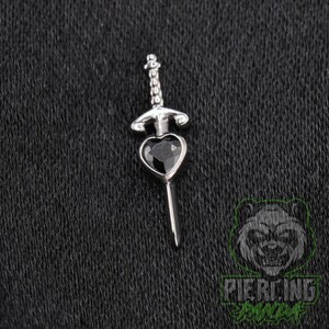 May include: A silver-toned metal body jewelry piece shaped like a sword with a heart-shaped black gemstone set in the center of the hilt. The sword features a detailed handle and a pointed blade, designed as a decorative piercing accessory.