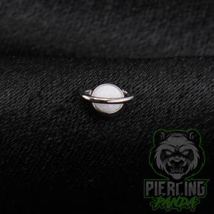 May include: A circular silver-toned captive bead ring featuring a smooth, white, opaque spherical center stone. The jewelry piece is centered and displays a polished metallic finish on the circular band that secures the round white bead in place.