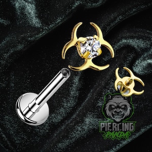 May include: A collection of body jewelry, including a silver labret stud and two gold-colored pieces with a biohazard symbol design. One piece features a clear, sparkling gem. The image is set against a dark, textured background.