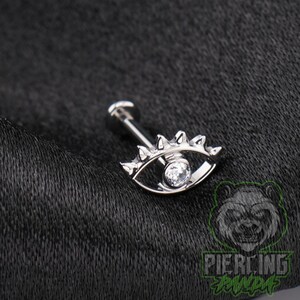 May include: A silver-toned flat back labret piercing featuring an eye-shaped charm. The charm has a central clear round gemstone as the pupil, surrounded by a metallic frame with small pointed spikes along the top edge of the eye shape.