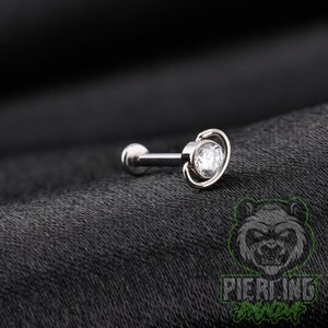May include: A silver-toned labret stud body jewelry piece featuring a circular clear gemstone set in a bezel, surrounded by a thin, smooth metal ring that encircles the stone, attached to a straight post with a flat circular back disc.