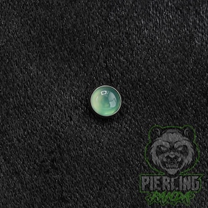May include: A circular body jewelry piece featuring a smooth, convex, light green stone cabochon set within a dark metal frame. The piece has a polished, reflective surface that catches light, creating a subtle sheen across the rounded top.