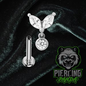 May include: A silver-colored lip piercing with a decorative top featuring two marquise-cut clear gems and a dangling round clear gem. The piercing post is also silver. The image includes a logo with a panda and the text "Piercing Panda."