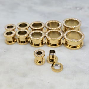 May include: A collection of gold-colored ear gauges with a sparkling crystal border. The gauges are in various sizes, with a few pieces disassembled. The jewelry is displayed on a light-colored surface.