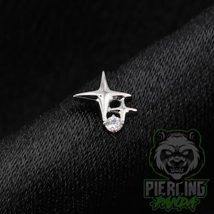 May include: A silver-toned metal body jewelry piece featuring a dual-star design with four-pointed rays. The jewelry is accented with a single, clear, round-cut gemstone set at the base of the smaller star, creating a bright, reflective finish.