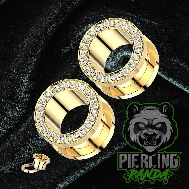 May include: Two gold-colored ear gauges with a ring of clear, sparkling stones around the outer edge. A smaller gauge is also visible. The gauges are on a dark green velvet-like surface. The logo "Piercing Panda" is in the bottom right corner.