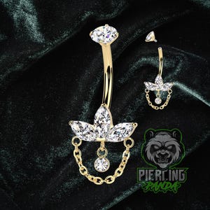 Peut inclure: Anneau de nombril doré avec un motif floral et une chaîne pendante. La bague est ornée de pierres précieuses claires et étincelantes. Une boucle d'oreille assortie plus petite est également présente. Le fond est en velours vert foncé.