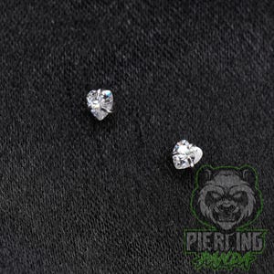 May include: Two silver-toned stud earrings, each featuring a clear, heart-shaped faceted gemstone held in place by small metal prongs. The earrings are positioned separately against a dark, textured background, showcasing their reflective surfaces.