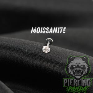 May include: A silver-toned labret piercing stud featuring a round, clear, faceted moissanite gemstone set in a four-prong metal setting. The word MOISSANITE is printed in white above the jewelry, and a green panda logo is visible below.