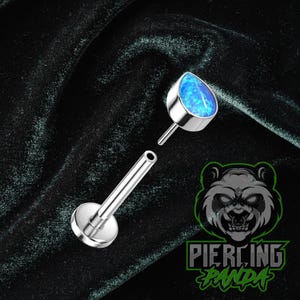Può includere: Un piercing per labbra color argento con una pietra di opale blu a forma di goccia. Il piercing è smontato, mostrando il perno e la base a disco piatto. Lo sfondo è un tessuto di velluto verde scuro.