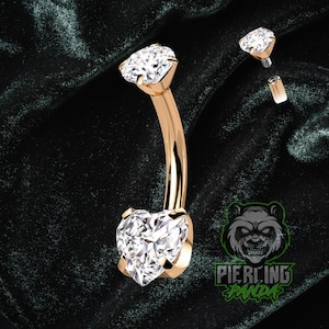 Pode incluir: Um piercing no umbigo dourado com um design de barra curva. O anel apresenta uma grande pedra preciosa transparente em forma de coração na parte inferior e uma pedra preciosa redonda transparente menor na parte superior. Componentes adicionais são visíveis.