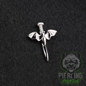 May include: A silver-toned metal body jewelry piece shaped like a dagger with bat wings extending from the hilt. The design features a central pointed blade, a ribbed handle, and two symmetrical wings. The surface is polished and reflective.