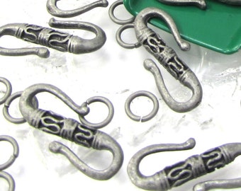 May include: An assortment of antique silver-colored S-shaped clasps and small jump rings. The clasps have an ornate, engraved design. Some clasps are in a green scoop, while others are scattered on a white surface. Ideal for jewelry making.