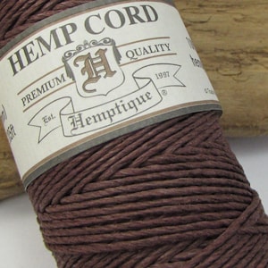Może przedstawiać: Szpula ciemnobrązowego sznurka konopnego z etykietą z napisem "Hemptique Hemp Cord". Etykieta zawiera również słowa "Premium Quality" i "Est. 1997". Sznurek jest ciasno nawinięty i ma około 45,72 metrów długości.