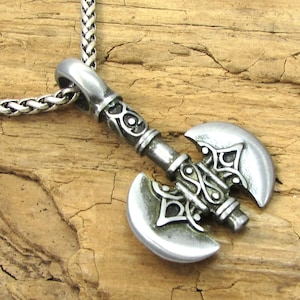 May include: A silver axe pendant with intricate details and a braided chain necklace. The axe has a double-headed blade with a decorative handle.