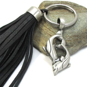 May include: A silver-toned dolphin charm keychain with a black leather tassel. The keychain features a ring and a charm depicting two dolphins. The tassel is made of black leather strips and has a silver-toned metal cap.