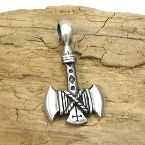 May include: A silver pendant in the shape of a double-headed axe with a rope design and a cross on the bottom. The pendant is on a wooden surface.